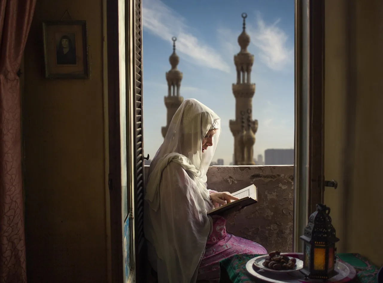 A woman in a white hijab reading the Quran beside a window